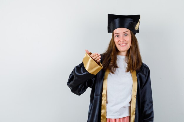 Graduada femenina levantando la mano para mostrar algo en traje académico y mirando alegre, vista frontal.