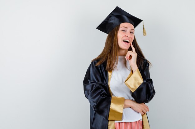 Graduada femenina manteniendo el dedo en la mejilla en traje académico y mirando alegre, vista frontal.