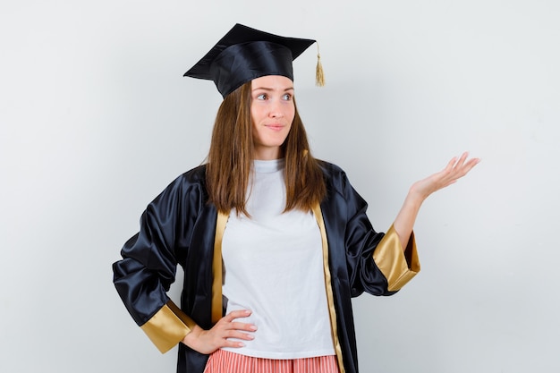 Graduada femenina mostrando gesto de bienvenida en uniforme, ropa casual y mirando enfocado. vista frontal.