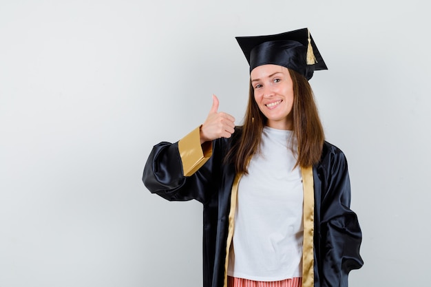 Graduada femenina mostrando el pulgar hacia arriba en traje académico y mirando alegre, vista frontal.