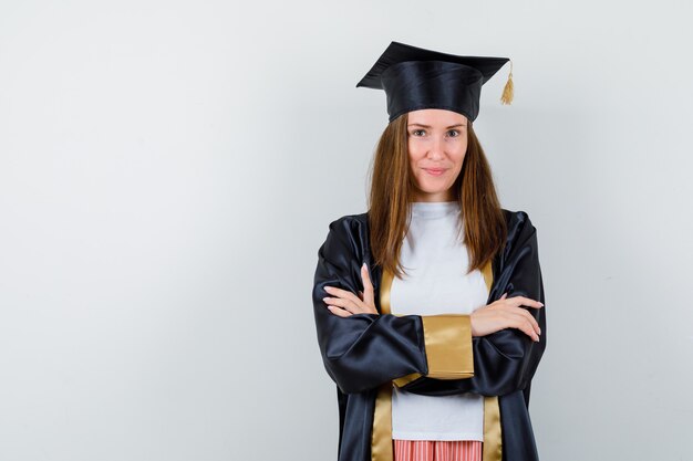 Graduada femenina de pie con los brazos cruzados en uniforme, ropa casual y con aspecto orgulloso. vista frontal.