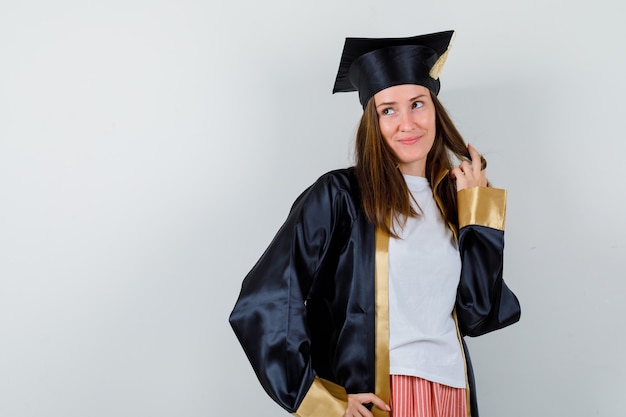 Foto gratuita graduada femenina posando mientras sostiene su mechón en uniforme, ropa casual y luciendo soñadora, vista frontal.