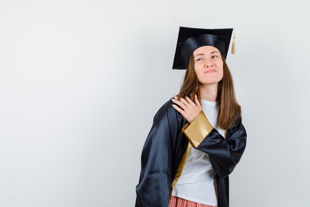 Graduada femenina que sufre de dolor en el hombro en uniforme, ropa casual y con aspecto incómodo, vista frontal.