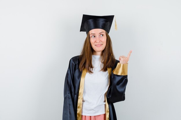 Graduada femenina en traje académico apuntando a la esquina superior derecha y mirando esperanzada, vista frontal.