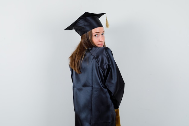 Graduada femenina en traje académico mirando a la cámara por encima del hombro y luciendo linda, vista posterior.