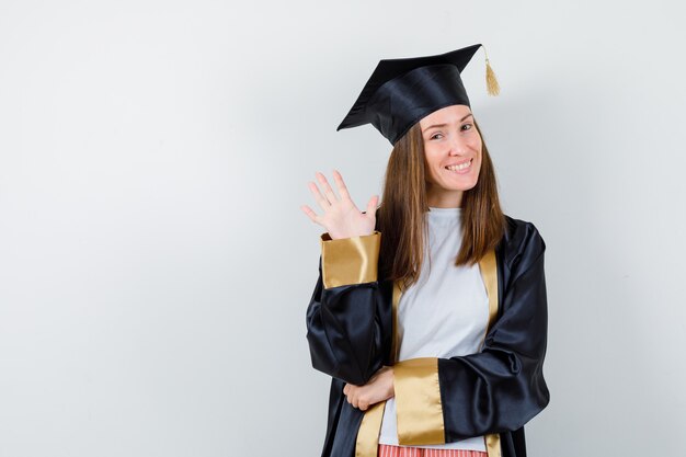 Graduada femenina en uniforme, ropa casual agitando la mano para decir adiós y mirando alegre, vista frontal.