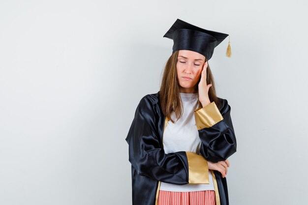 Graduada femenina en uniforme, ropa casual con dolor de cabeza y luciendo agotada, vista frontal.