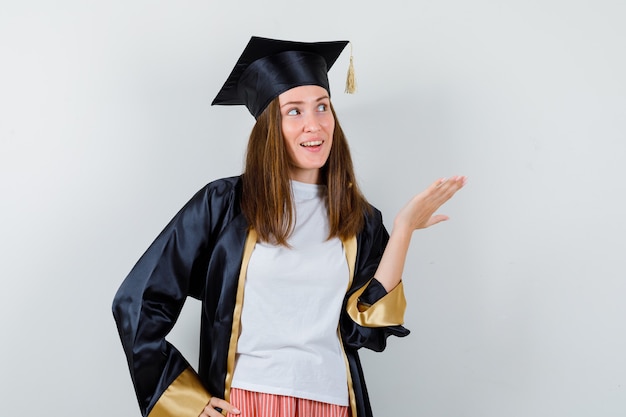 Graduada femenina en uniforme, ropa casual mostrando algo mientras mira hacia arriba y se ve alegre, vista frontal.