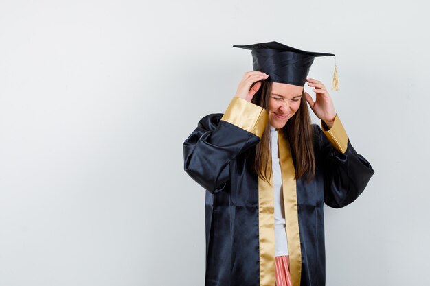 Graduada joven cogidos de la mano cerca de la cabeza en traje académico y mirando molesto, vista frontal.