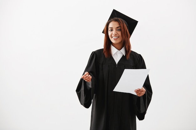 Graduado alegre feliz de la mujer que sonríe llevando a cabo la prueba sobre la superficie blanca