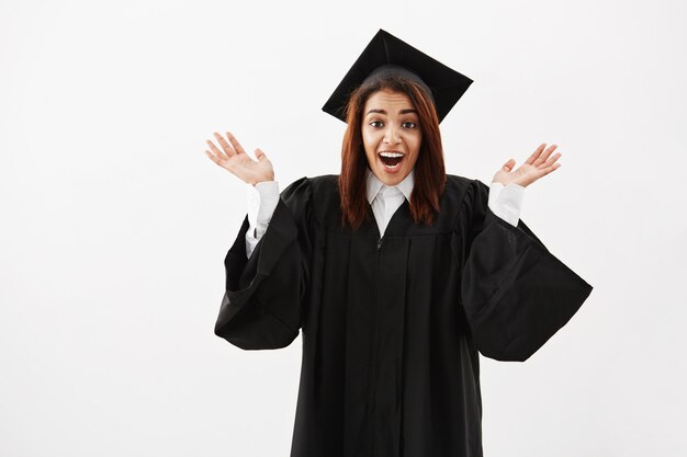 Graduado feliz de la mujer sorprendida que gesticula mirando la cámara sobre la superficie blanca