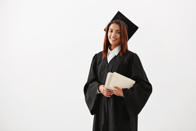 Graduado femenino africano alegre hermoso que sonríe sosteniendo los libros. Copia espacio