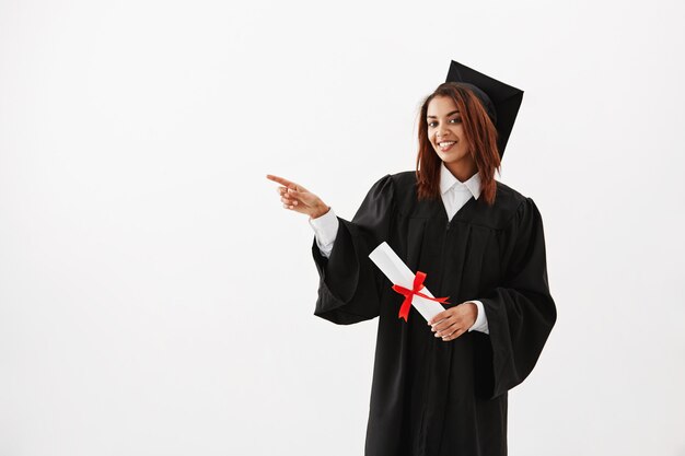 Graduado femenino africano hermoso que sonríe señalando el dedo en lado.