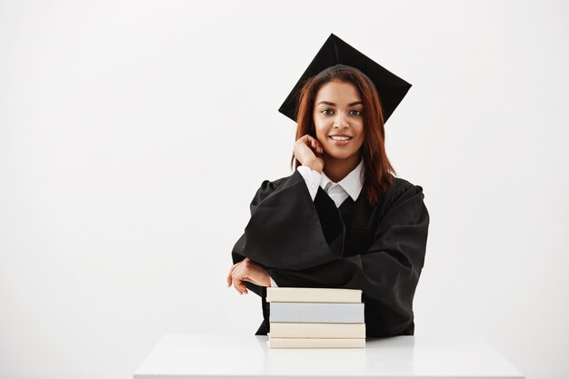 Graduado femenino en gorra y manto sonriendo sentado con libros sobre superficie blanca