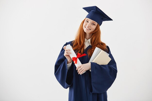 Graduado femenino hermoso del pelirrojo que sonríe sosteniendo los libros y el diploma.