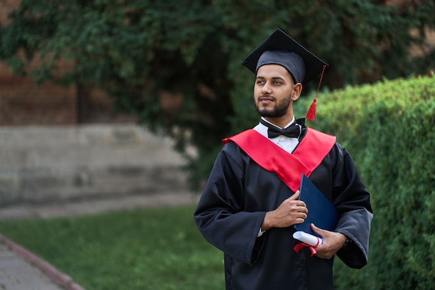 Graduado indio en bata de graduación con diploma en espacio de copia del campus universitario.