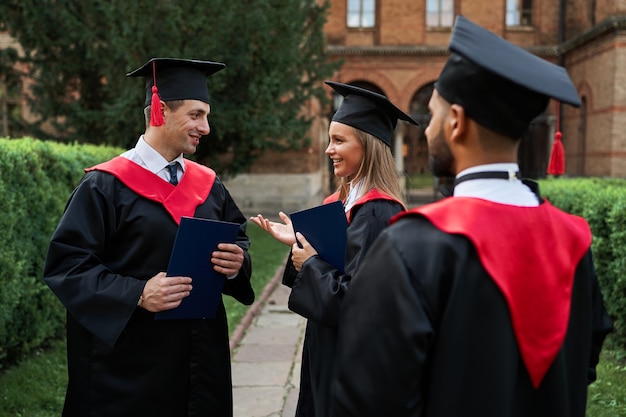 Graduado de mujeres y hombres multinacionales hablando con batas de graduación en el campus universitario.