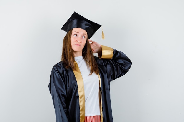 Graduado de sexo femenino joven que lleva a cabo la mano detrás de la cabeza en traje académico y que mira vacilante. vista frontal.