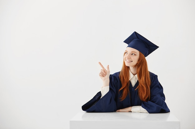 Graduado universitario femenino joven en el casquillo académico que se sienta en la tabla que sonríe señalando a la izquierda. Futuro abogado o ingeniero mostrando una idea.