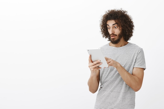 Gráfico de empresa subido inesperadamente. Estudiante masculino guapo sorprendido y complacido con cabello rizado y barba, dejando caer la mandíbula mientras se desplaza por las noticias en la tableta digital