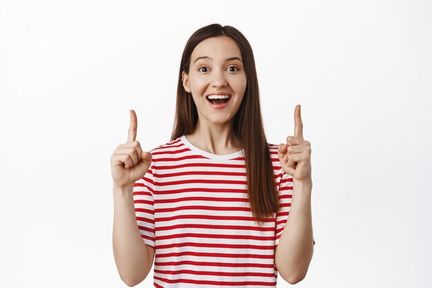 Gran anuncio en la parte superior Emocionada niña sonriente señalando con el dedo feliz mostrando el logotipo de anuncio de venta o pancarta hacia arriba de pie contra el fondo blanco
