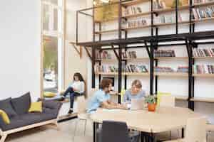 Foto gratuita gran biblioteca moderna en la mañana. dos personas sentadas, mirando en el monitor del portátil hablando sobre el proyecto de inicio. chica sentada en el alféizar de la ventana de lectura en el teléfono inteligente pasar tiempo antes de estudiar.