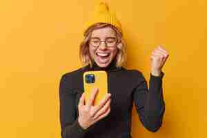 Foto gratuita gran concepto de suerte una mujer joven de cabello rubio muy contenta aprieta el puño exclama de alegría exclama con emoción usa sombrero y cuello alto negro aislado sobre fondo amarillo ganó la lotería en línea