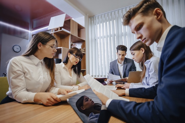 Un gran equipo de personas está trabajando en una mesa para computadoras portátiles, tabletas y papeles, en el fondo un gran televisor en una pared de madera