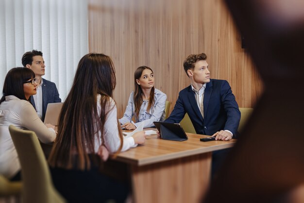 Un gran equipo de personas está trabajando en una mesa para computadoras portátiles, tabletas y papeles.