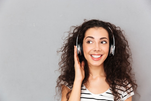 Gran imagen de mujer caucásica rizada en camiseta a rayas en auriculares disfrutando de la música a través de un dispositivo moderno mientras descansa