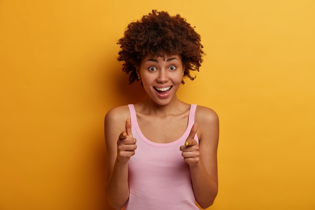 Gran oferta para ti. Alegre mujer de pelo rizado muestra un signo de pistola, se ríe positivamente, expresa su elección y elige a alguien, viste una camisa informal, aislada en una pared amarilla. Hey te recordaba