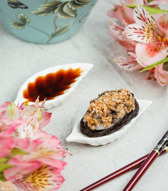 Gran pieza de sushi nori con salsa de soja dentro de un plato en forma de hoja blanca con flores alrededor.