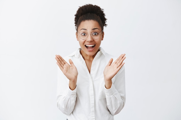 Gran plan, adoro. Retrato de mujer feliz asombrada y entusiasta con gafas y camisa blanca, aplaudiendo en las palmas y sonriendo con expresión emocionada, siendo levantada de una gran idea sobre una pared gris