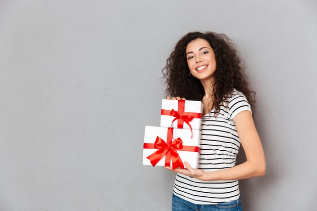 Grandes emociones de mujer joven en camiseta a rayas con dos cajas envueltas en regalos con lazos rojos mientras está de pie sobre la pared gris