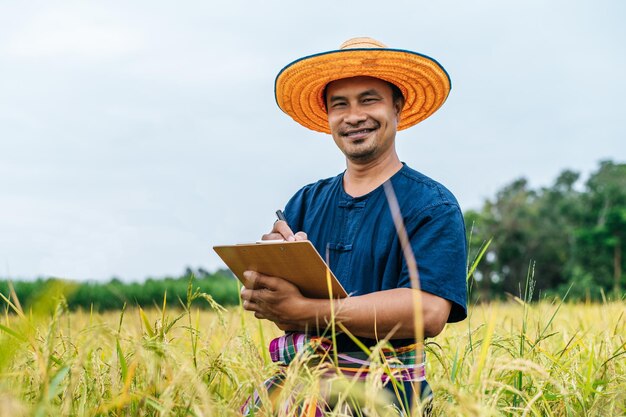 Granjero asiático de mediana edad hombre vestido con sombrero de paja escribir en el portapapeles en el campo de arroz con una sonrisa durante la conservación de datos