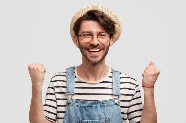 Un granjero positivo de buen aspecto levanta los puños cerrados, se siente satisfecho y emocionado, logra un gran éxito en la esfera agrícola, usa un mono informal, suéter a rayas, sombrero de paja, tiene una amplia sonrisa