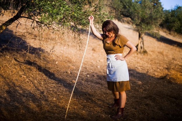 Foto gratuita granjero de sexo femenino agotado que se coloca con el palillo en un campo de la huerta de olivo