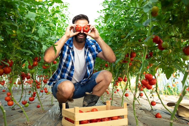 Granjero trabajador haciendo caras tontas y divertidas con verduras de tomate en el jardín