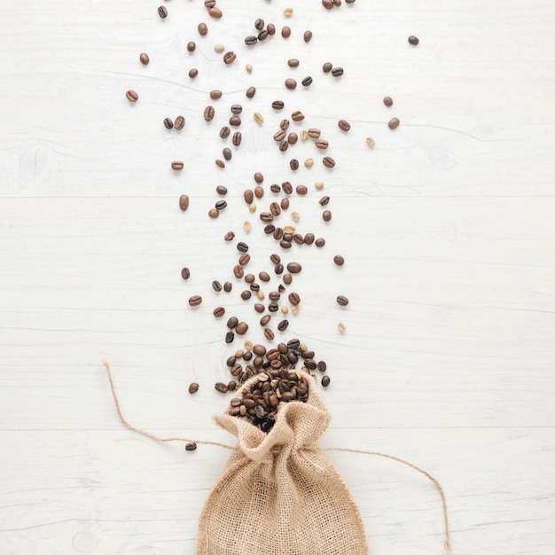 Foto gratuita granos de café crudos y tostados que caen de un pequeño saco en el escritorio