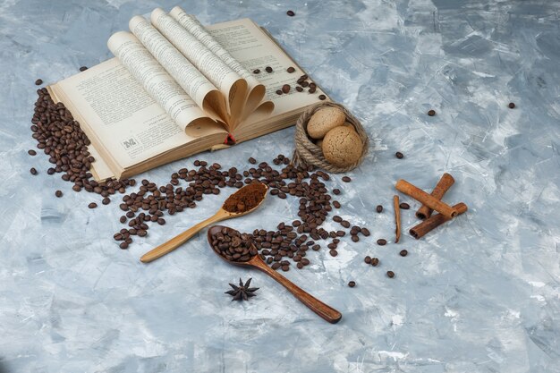 Granos de café en una cuchara de madera con café molido, libro, especias, galletas vista de ángulo alto sobre un fondo gris sucio