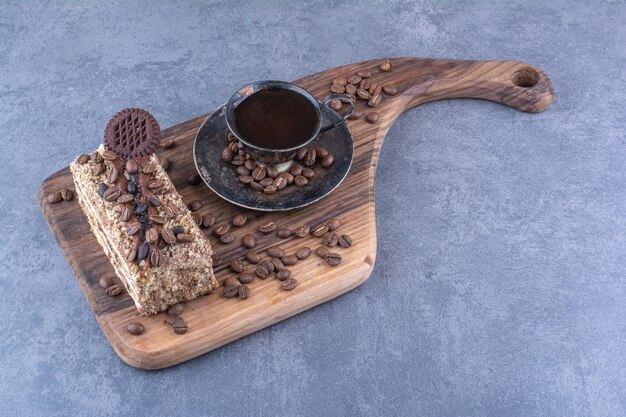 Granos de café esparcidos alrededor de una taza de café y una rebanada de pastel sobre una tabla de madera sobre una superficie de mármol