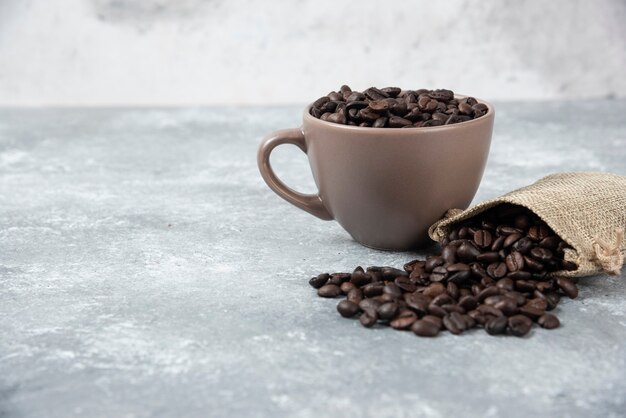 Granos de café tostados en saco de arpillera y en taza sobre mármol.