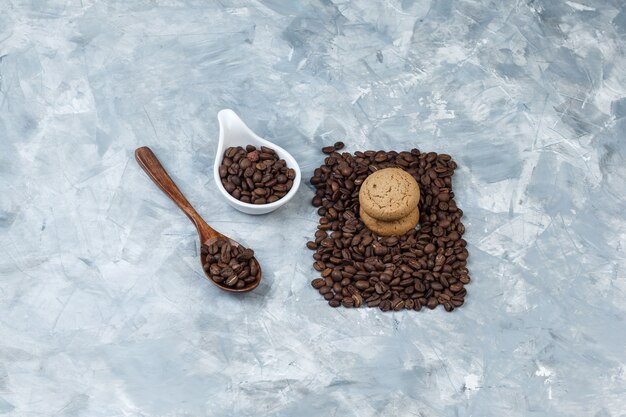 Granos de café de vista de ángulo alto en cuchara de madera, jarra de porcelana blanca con galletas sobre fondo de mármol azul claro. horizontal