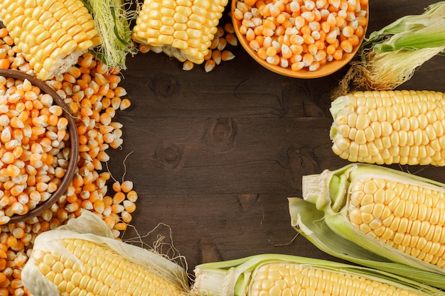 Granos de maíz con mazorcas en cuchara de madera y plato en la mesa de madera, endecha plana.