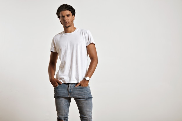 Grave atlético joven modelo afroamericano con las manos en los bolsillos de sus jeans ajustados con una camiseta blanca