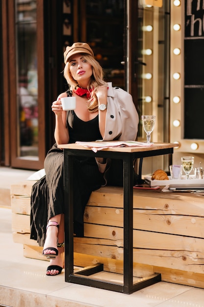 Grave chica rubia con zapatos negros enfriándose en un café al aire libre y disfrutando del té. Mujer joven atractiva viste sandalias con estilo y sombrero marrón mirando a otro lado, sosteniendo una taza de café.