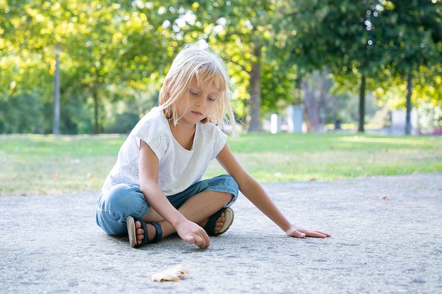 Grave dulce niña rubia sentada y dibujando con coloridos trozos de tizas. Copie el espacio. Concepto de infancia y creatividad.