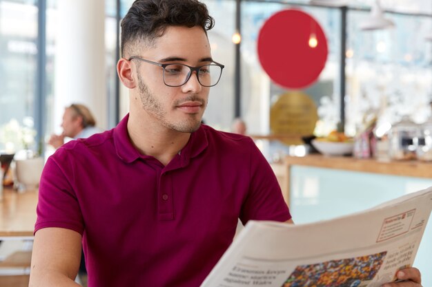 Grave economista masculino sin afeitar lee atentamente noticias del periódico, centrado en el atrículo financiero, recibe prensa comercial, usa gafas y camiseta roja informal, modelos contra el interior del café