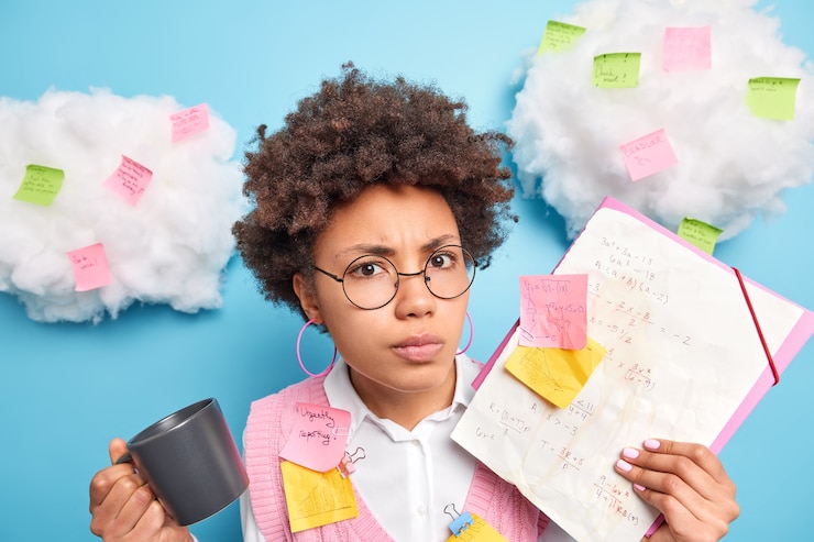 Foto gratuito grave, enojada, disgustada colegiala con cabello afro se prepara para el examen de matemáticas toma notas intenta recordar fórmulas, bebidas, café, usa anteojos redondos, posa contra adhesivos de pared azules alrededor