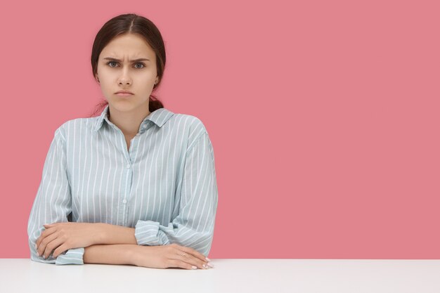 Grave estudiante disgustada vestida con camisa a rayas manteniendo los codos sobre el escritorio, frunciendo el ceño, enojada con una mala calificación injusta dada por un maestro estricto. Retrato aislado contra la pared rosa